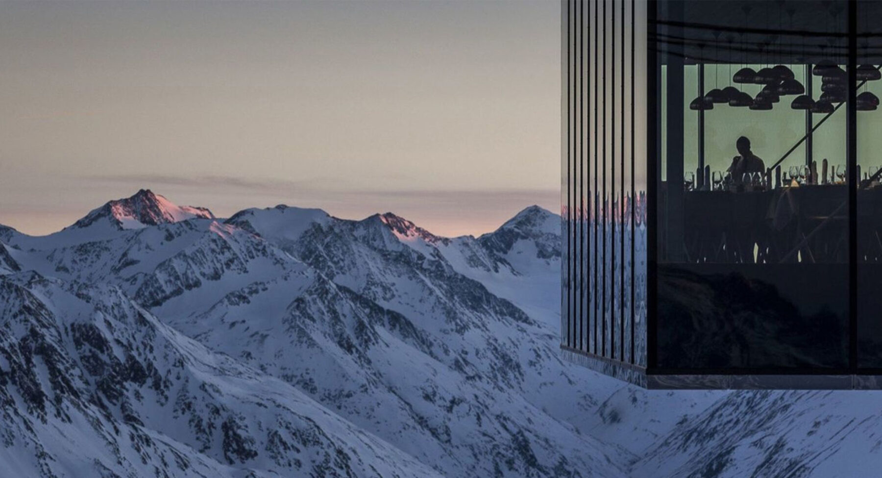 Glass-walled restaurant overlooking a sunset over snowy mountains