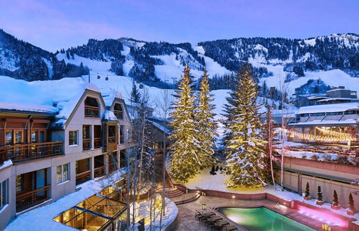 The exterior of the luxury ski hotel the Little Nell Hotel in Aspen with mountains in the background and trees and a swimming pool in the foreground